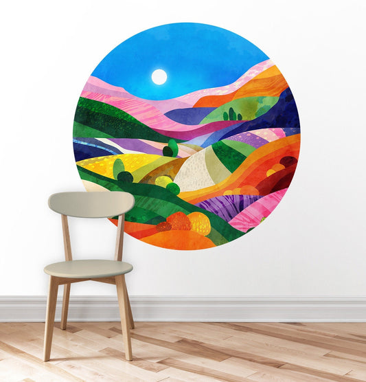 A colorful circular wall art print depicting an abstract landscape with rolling hills under a blue sky, shown on a white wall above a light wood floor with a beige chair in front.