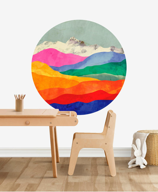 A large circular wall mural featuring a colorful watercolor mountain landscape with pink, green, blue, orange, yellow, and red layers