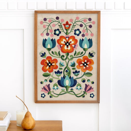 A framed folk art print featuring a colorful floral pattern on a cream background, hung on a white paneled wall above a wooden table with a pear and candle.