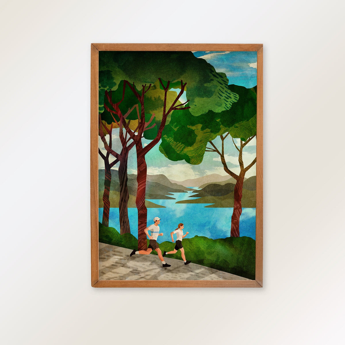 A framed art print depicting two runners jogging along a path by a blue lake, surrounded by green trees and hills, shown hanging on a light gray wall.