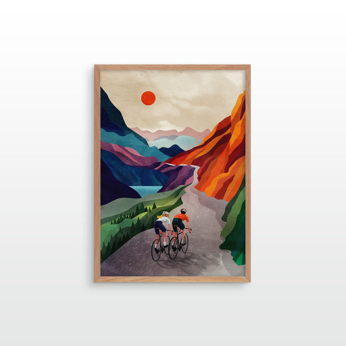 A framed art print featuring a stylized illustration of two cyclists riding along a winding mountain road, with colorful peaks, a red sun, and a light wood frame, shown against a plain white wall.