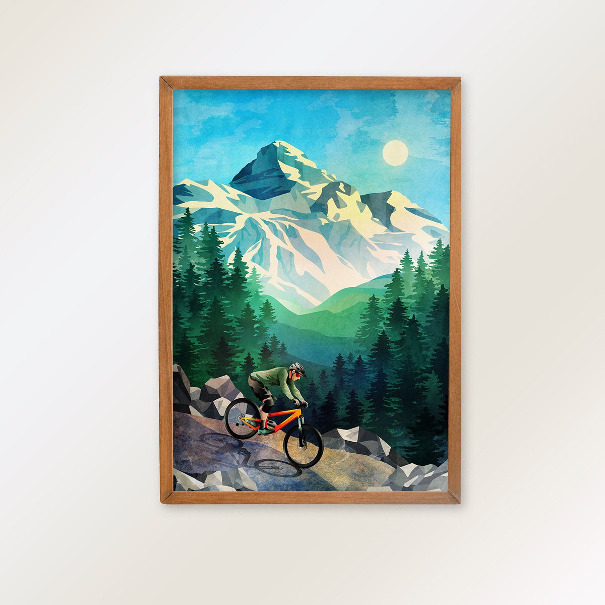 A framed art print depicting a mountain biker riding down a rocky trail with snowy mountains and pine trees in the background, hung on a plain white wall.