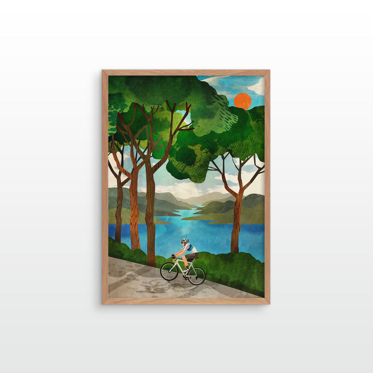 A framed art print featuring a stylized illustration of a cyclist riding along a path by a blue lake, surrounded by green trees and hills under a warm orange sun, shown against a plain white background.