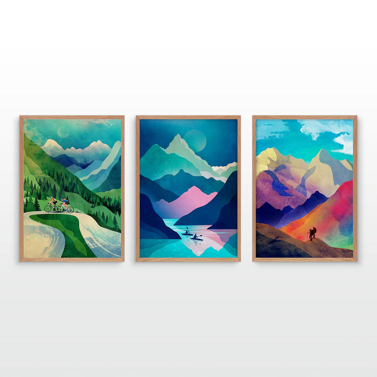 Three framed vertical art prints depicting colorful mountain landscapes, displayed side-by-side on a white wall. The left print shows a cyclist on a winding road through green and blue mountains.