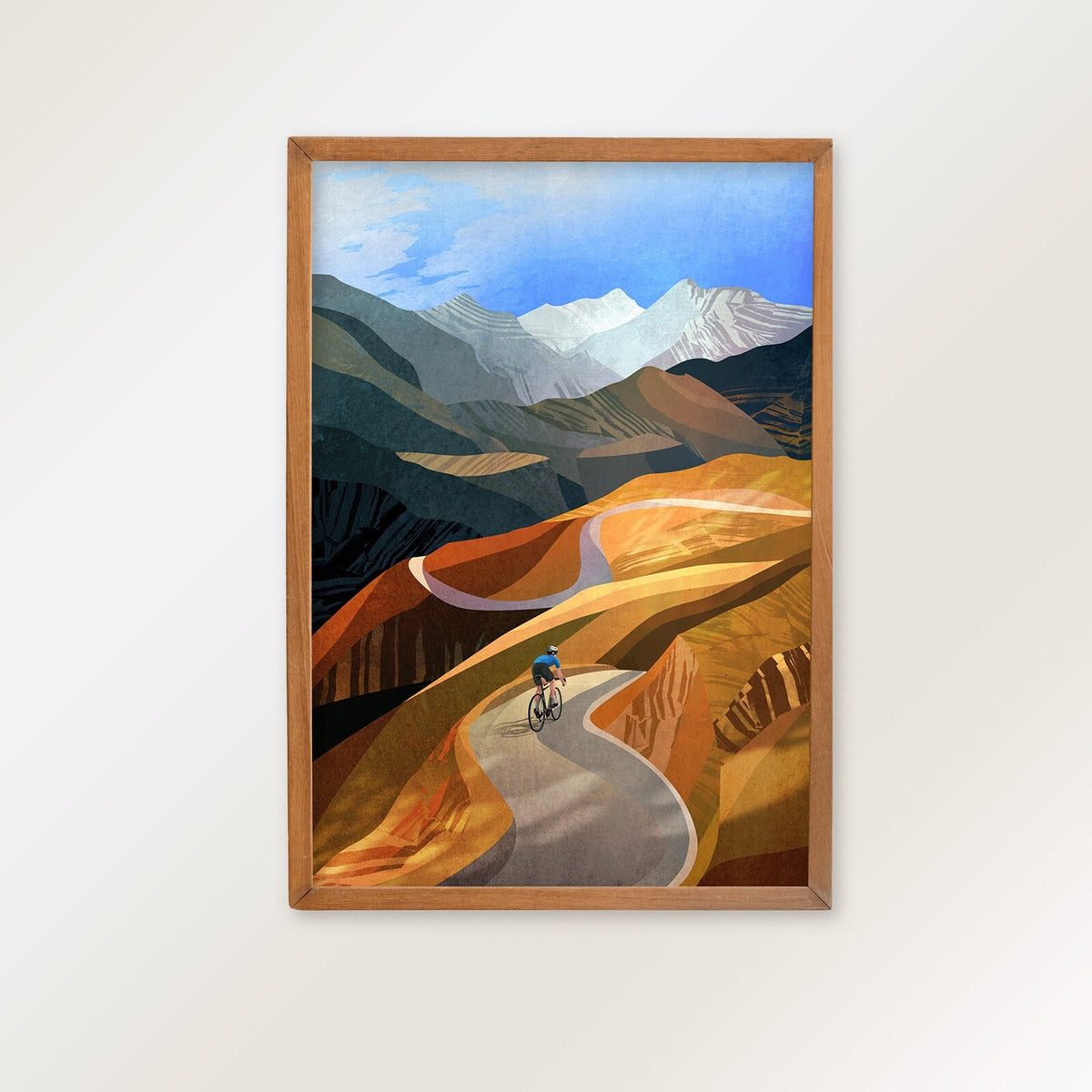 A framed art print depicting a stylized landscape with a cyclist on a winding road, set against mountains under a blue sky, hung on a plain white wall.