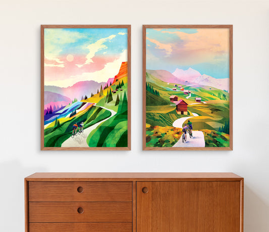 Two framed art prints depicting colorful mountain landscapes with cyclists, displayed side-by-side on a white wall above a wooden cabinet.