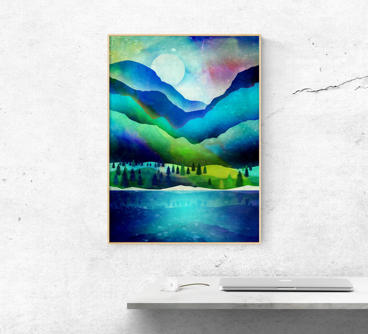 A framed abstract landscape art print featuring blue, green, and teal mountain and lake scenes, displayed on a light gray textured wall above a white shelf with a laptop and small object.