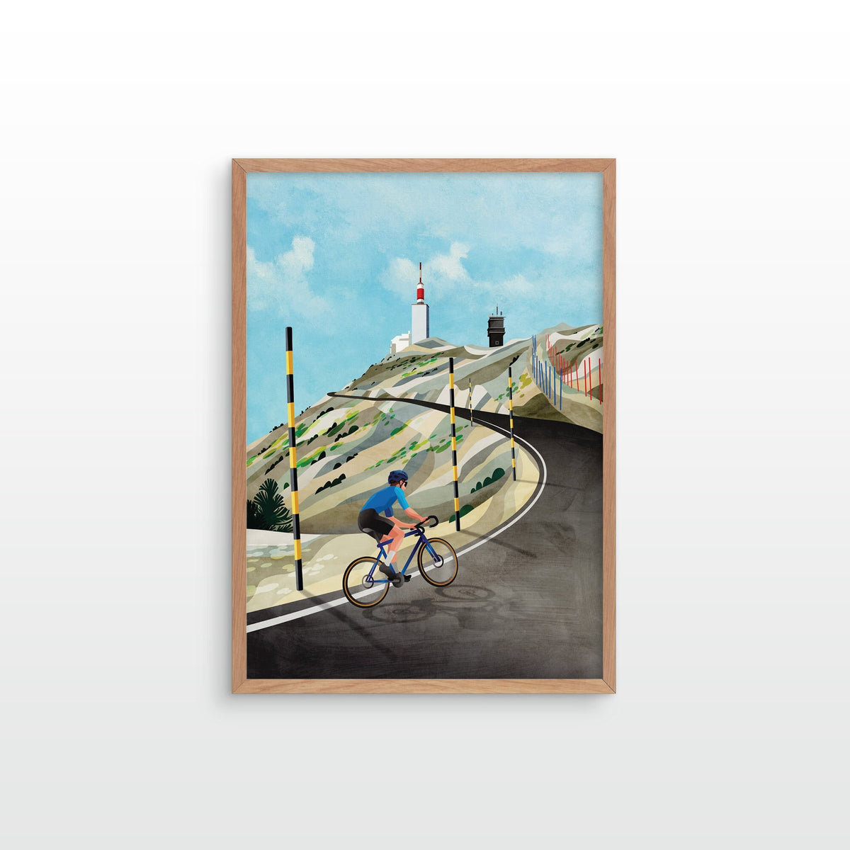 A framed art print featuring a stylized illustration of a cyclist on a winding road with a lighthouse in the background, set in a light wood frame against a white wall.