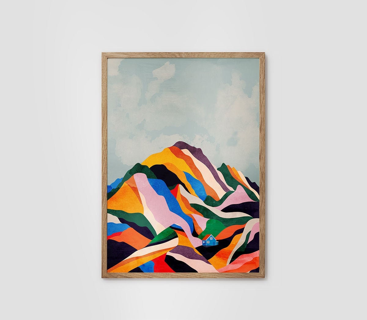 A framed art print featuring a colorful, abstract mountain landscape with geometric shapes in orange, yellow, green, blue, pink, and purple against a pale sky, set in a light wood frame on a neutral wall.