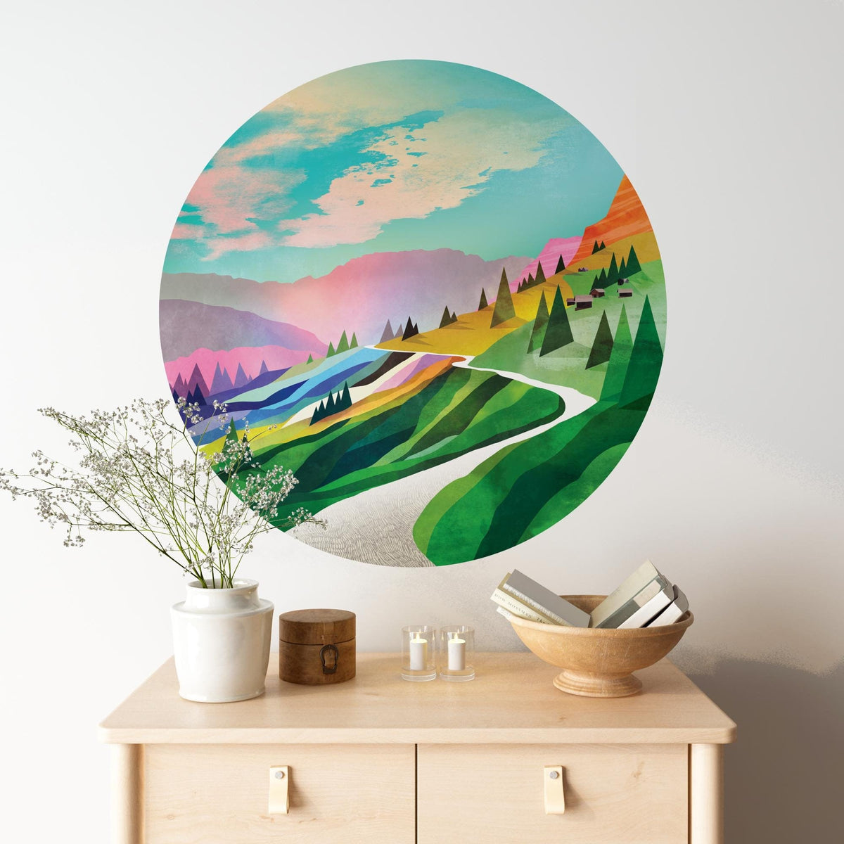 A large circular wall art print featuring a colorful stylized landscape with green hills, blue water, pink mountains, and a winding white road, displayed above a light wood console table with decorative objects.