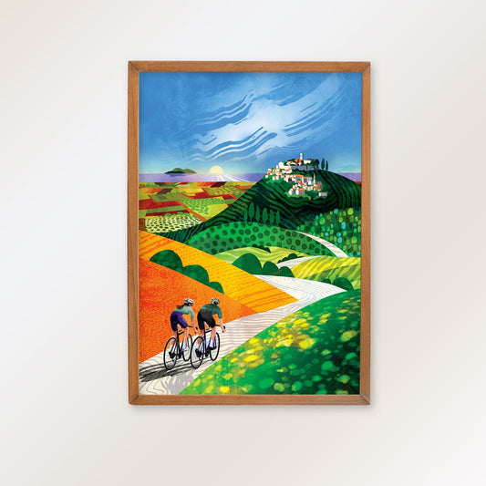 A framed art print featuring a colorful landscape illustration with two cyclists on a winding road, hanging on a plain white wall.