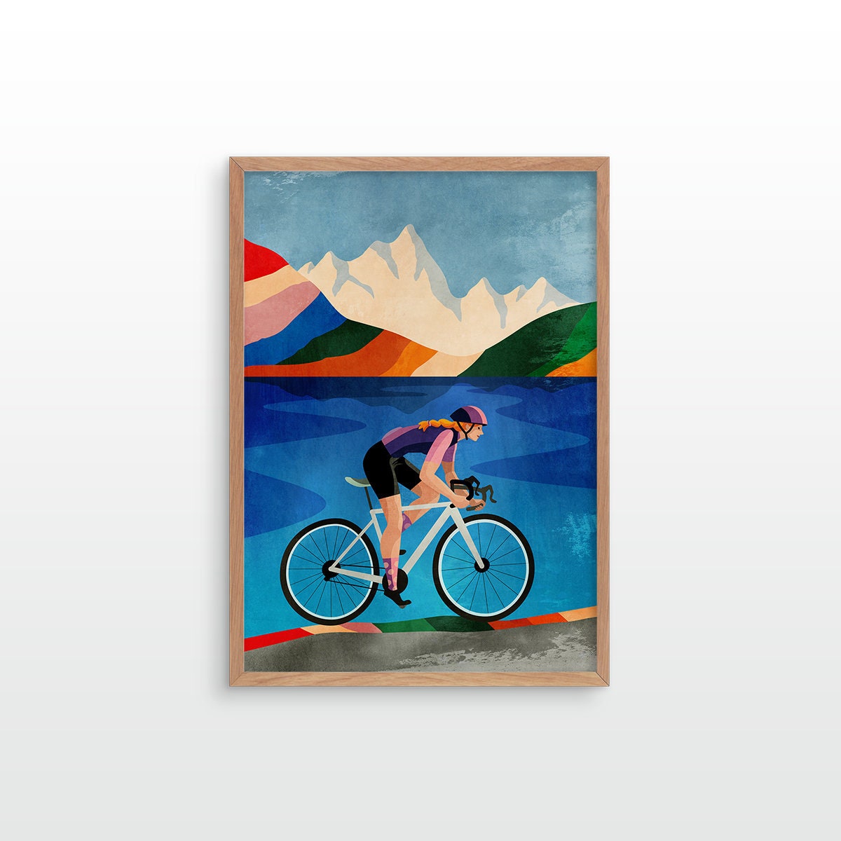 A framed art print featuring a stylized illustration of a cyclist riding a road bike against a backdrop of blue water, green hills, and white mountains, displayed on a light gray wall.