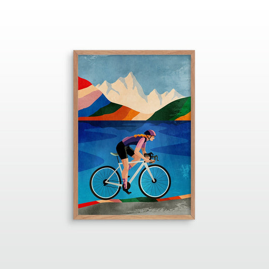 A framed art print featuring a stylized illustration of a cyclist riding a road bike against a backdrop of blue water, green hills, and white mountains, displayed on a light gray wall.