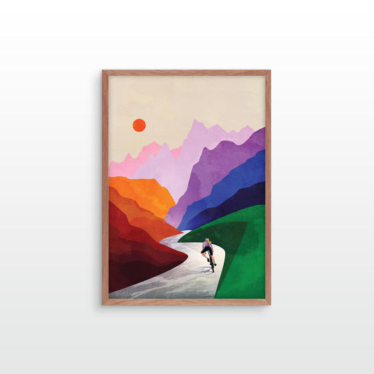 A framed art print featuring a stylized landscape with layered mountains in purple, pink, orange, and red, a green foreground, a winding light gray path, and a small cyclist. 
