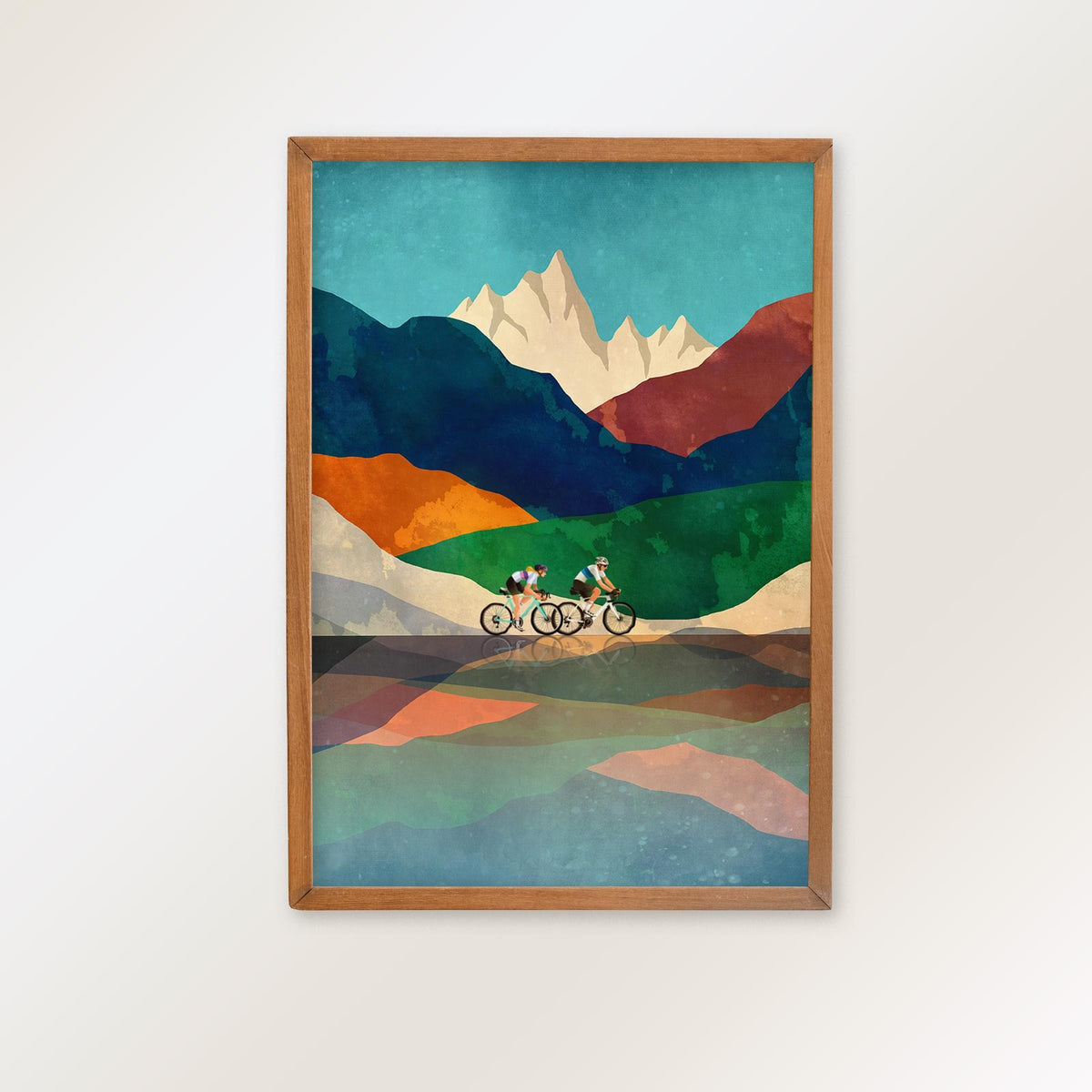 A framed art print featuring a stylized mountain landscape with two cyclists on a road, hung on a plain white wall.