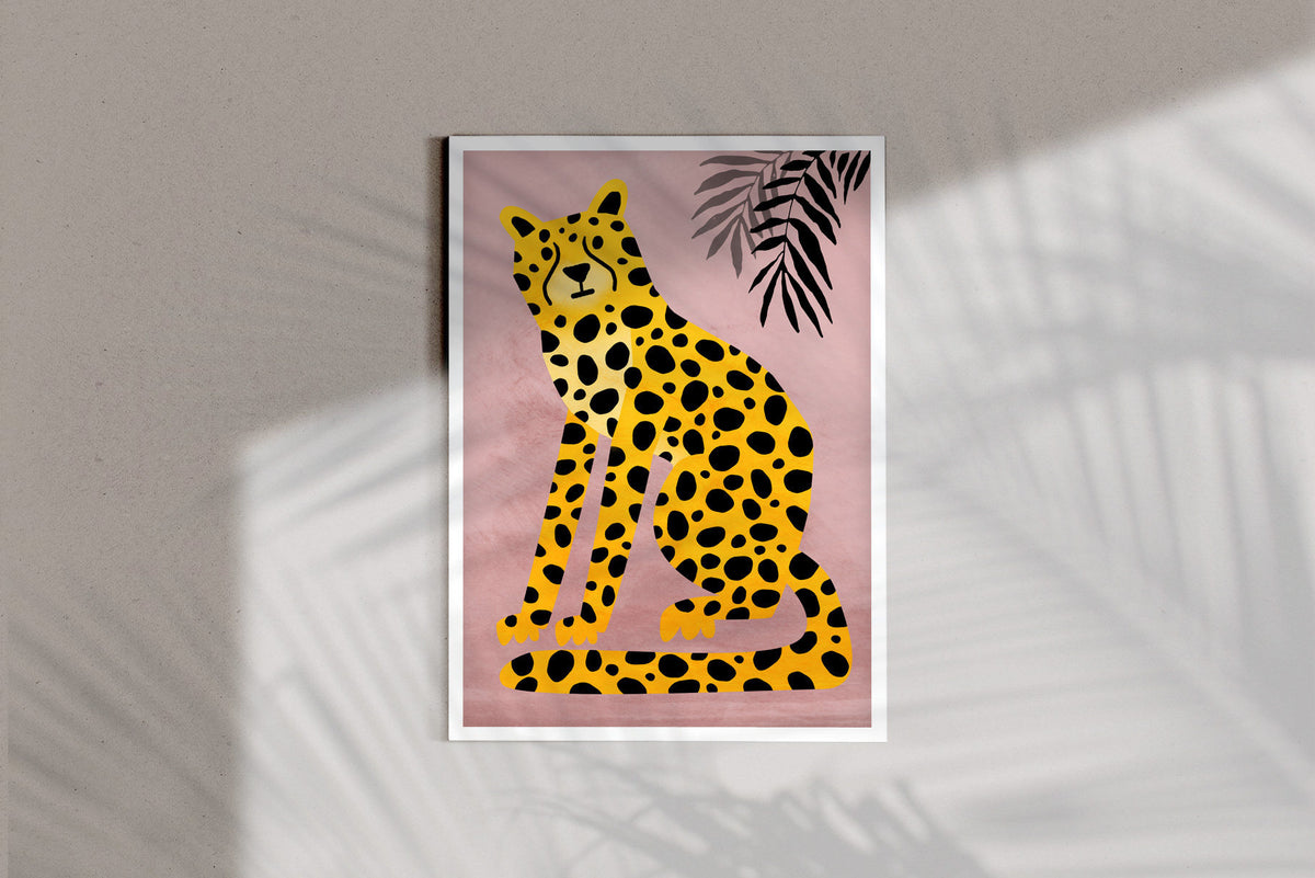 A framed art print featuring a stylized yellow cheetah with black spots on a dusty pink background, with a black palm leaf in the upper right. The print is displayed on a neutral beige wall with soft palm leaf shadows cast across it.