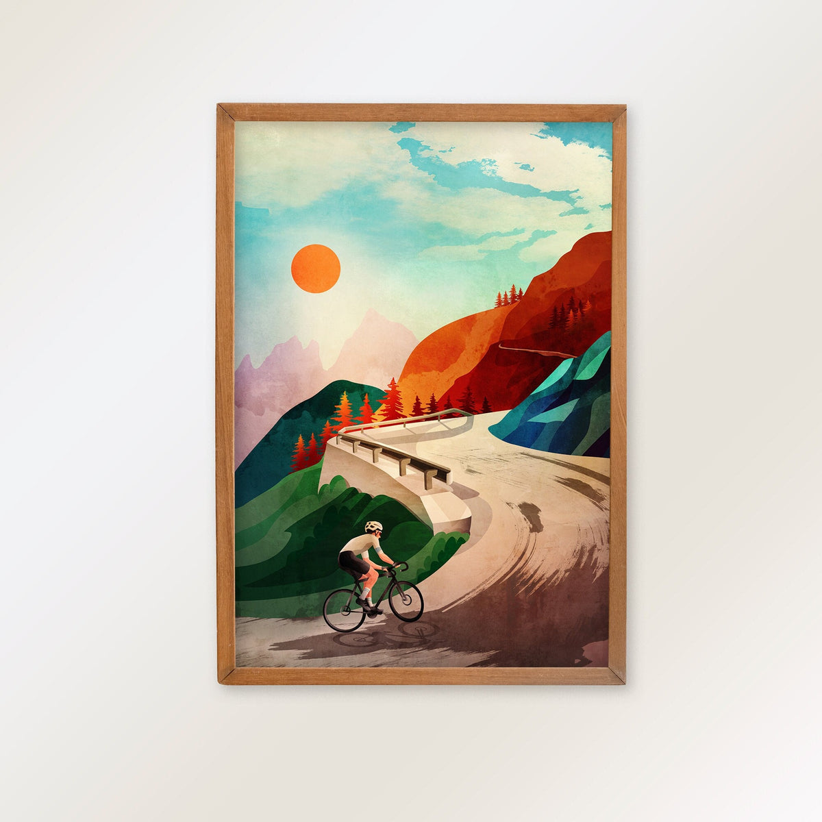 A framed art print featuring a retro-style illustration of a cyclist on a winding road through a mountain landscape under a bright sun, hung on a plain white wall.