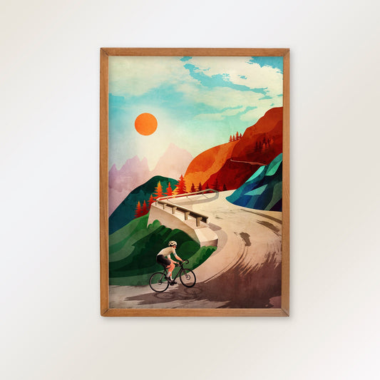 A framed art print featuring a retro-style illustration of a cyclist on a winding road through a mountain landscape under a bright sun, hung on a plain white wall.