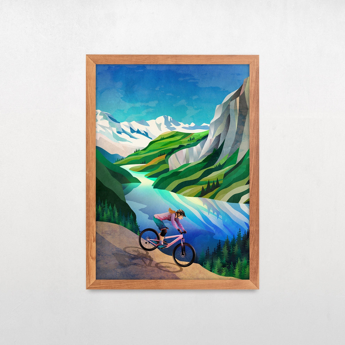 A framed art print featuring a stylized illustration of a woman riding a pink mountain bike through a scenic green valley with blue water and snowy mountains, displayed on a light gray wall.