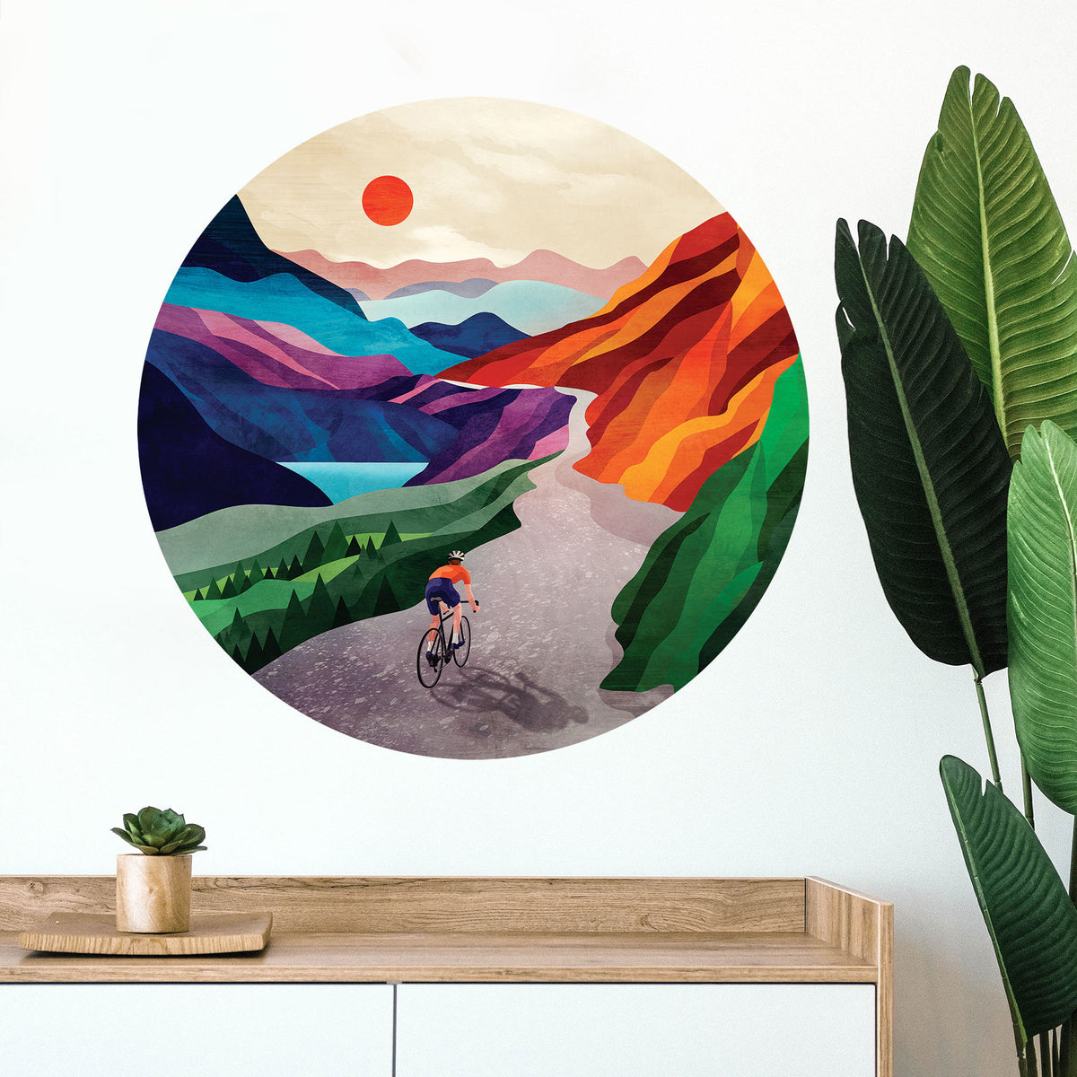 A circular wall art print featuring a stylized mountain landscape with a cyclist on a road, displayed on a white wall above a wooden console table with a small plant and a large green leafy plant nearby.