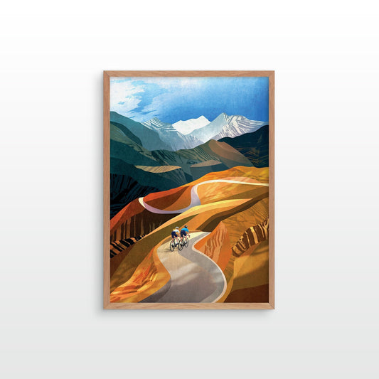 A framed art print depicting a stylized mountain landscape with a winding road and two cyclists, shown hanging on a light gray wall.