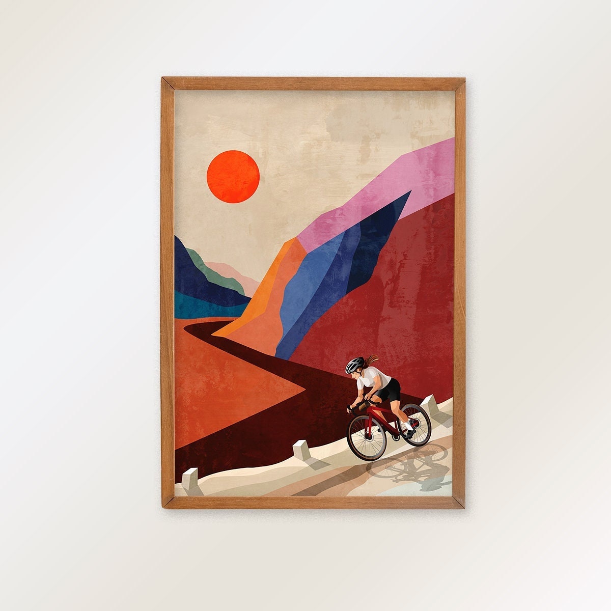 A framed art print featuring a stylized illustration of a cyclist riding along a winding road through geometric mountains under a large orange sun, hung on a plain light wall.