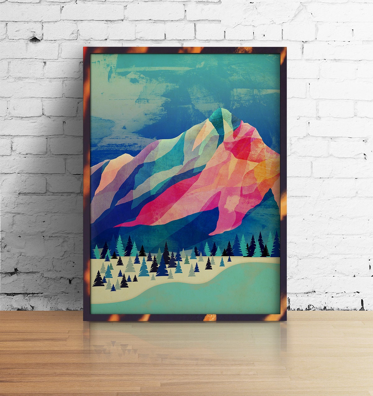 A framed art print featuring a colorful geometric mountain landscape leans against a white brick wall on a light wood floor. The print has a tortoiseshell-style frame and displays a low-poly style mountain with pink, teal, blue, and yellow facets above a pine forest.