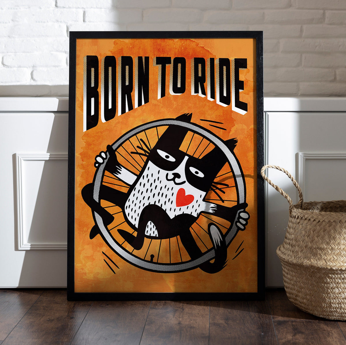A framed art print leaning against a white paneled wall on a dark wood floor, next to a woven basket. The print features a black and white cartoon cat riding a bicycle wheel against a textured orange background, with the text 'BORN TO RIDE' in bold black letters.
