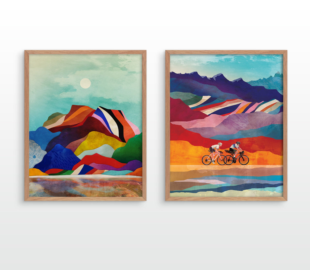 Two framed abstract landscape art prints hanging side by side on a white wall. The left print shows colorful mountain-like forms under a pale sky with a sun, and the right print shows two cyclists riding across a stylized landscape with mountains, stripes, and reflective water.