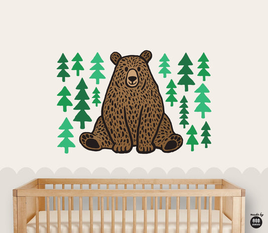 A wall decal featuring a brown bear and green pine trees, shown applied to a nursery wall above a wooden crib.