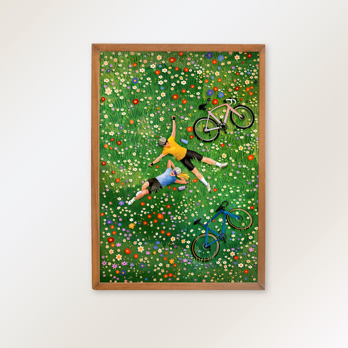 A framed art print depicting two people lying on a green flower-covered field with bicycles, shown hanging on a light gray wall.