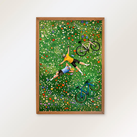 A framed art print depicting two people lying on a green flower-covered field with bicycles, shown hanging on a light gray wall.