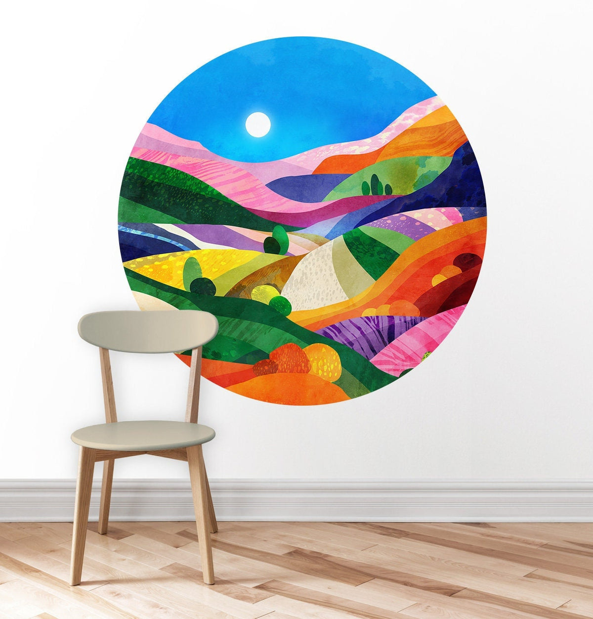 A colorful circular wall art print depicting an abstract landscape with rolling hills under a blue sky, shown on a white wall above a light wood floor with a beige chair in front.