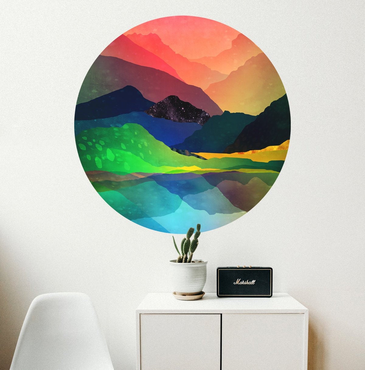 A large circular wall decal featuring a colorful watercolor mountain landscape with green fields, blue lakes, and a pink-orange sunset, displayed on a white wall above a small white cabinet with a potted cactus and a black Marshall speaker.