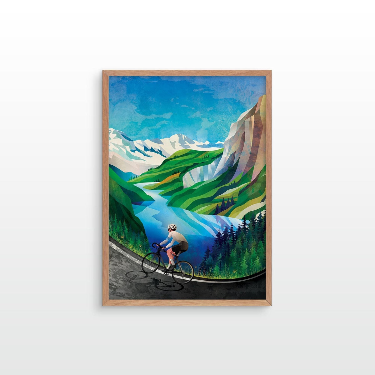 A framed art print depicting a cyclist riding along a winding road through a vibrant mountain landscape with green hills, blue sky, and snow-capped peaks, shown on a light gray wall.