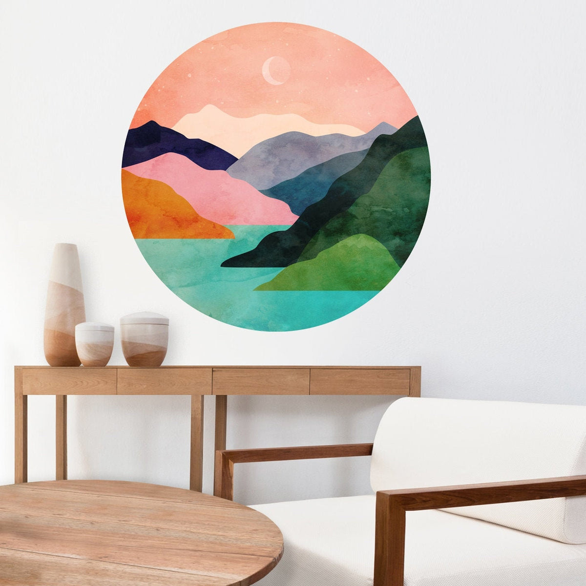 A circular watercolor-style wall art print showing layered mountains, green hills, turquoise water, and a peach sky with a crescent moon, displayed above a wooden console table in a modern living room setting.