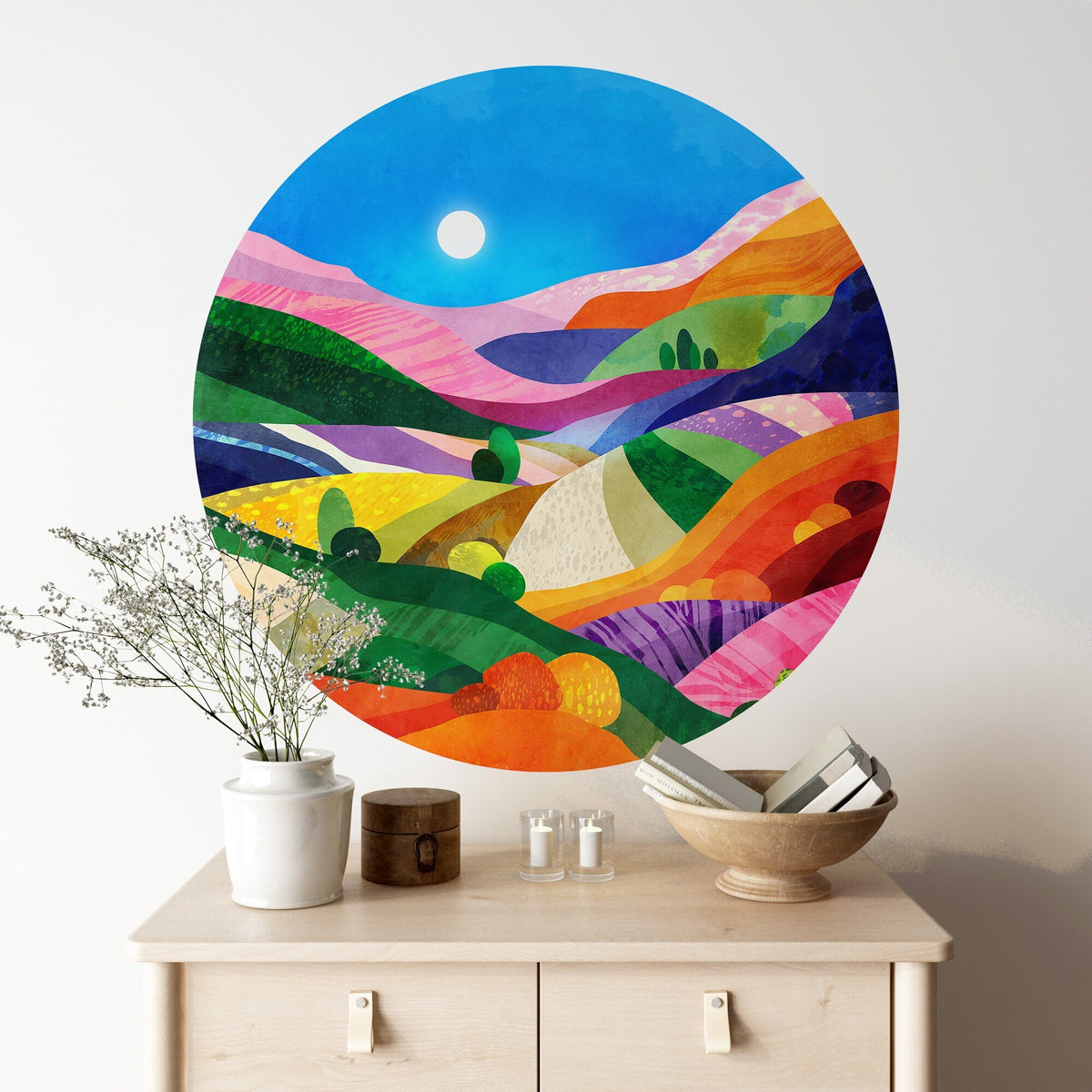 A colorful circular wall art print depicting an abstract landscape with rolling hills under a blue sky, shown on a white wall 