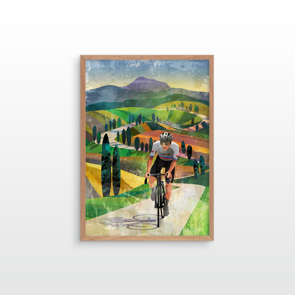 A framed art print depicting a cyclist riding along a scenic green and yellow path with mountains in the background, mounted on a light gray wall.