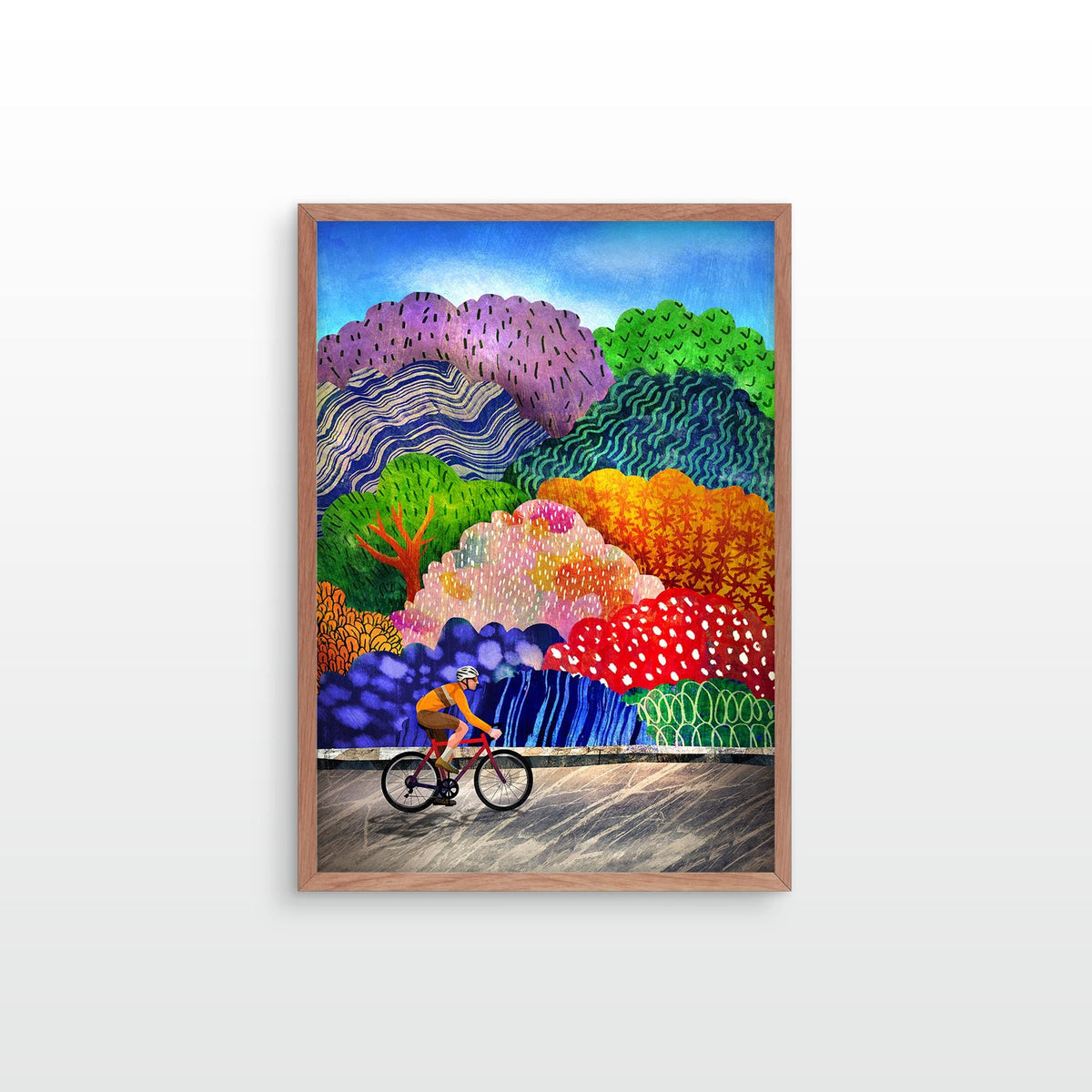 A framed colorful landscape art print featuring a cyclist on a road with stylized hills and trees in purple, green, orange, red, and blue, shown hanging against a light gray wall.