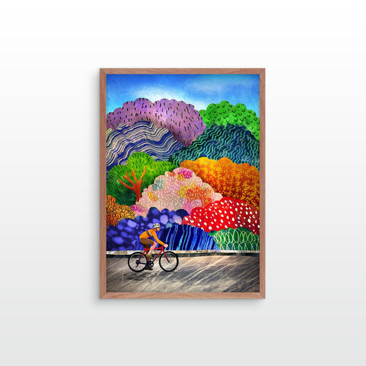 A framed colorful landscape art print featuring a cyclist on a road with stylized hills and trees in purple, green, orange, red, and blue, shown hanging against a light gray wall.