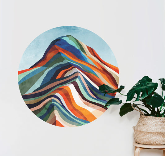 A circular wall art print featuring an abstract mountain design with layered wavy stripes in blue, orange, red, green, and cream, displayed on a white wall next to a potted green plant.