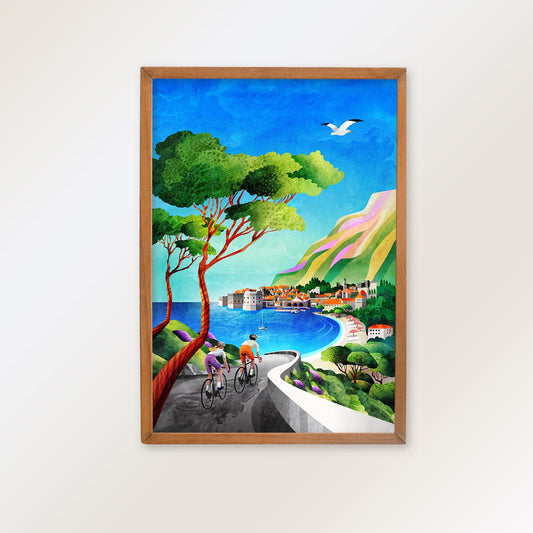 A framed art print depicting a colorful coastal village scene with cyclists, a seagull, a winding road, and a blue sea under a bright sky, hung on a light beige wall.