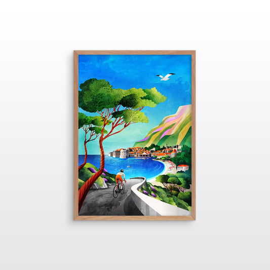 A framed art print featuring a colorful coastal landscape with a cyclist on a winding road, a large green tree, blue sea, pastel hills, and a bird in the sky, shown hanging on a light gray wall.