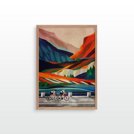 A framed art print featuring a colorful, cubist-style landscape with two cyclists on a road in the foreground, displayed against a clean white wall.